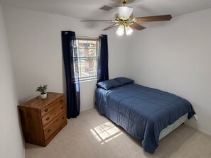 A bedroom with a bed, a wooden dresser, a ceiling fan, and a window with curtains.