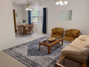 Una sala de estar con un sofá, una mesa de centro, un tapete estampado, un área de comedor con sillas y una ventana con cortinas.