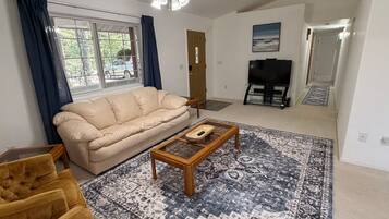 Un salon avec un canapé beige, un tapis à motifs, une table basse et un ventilateur de plafond.