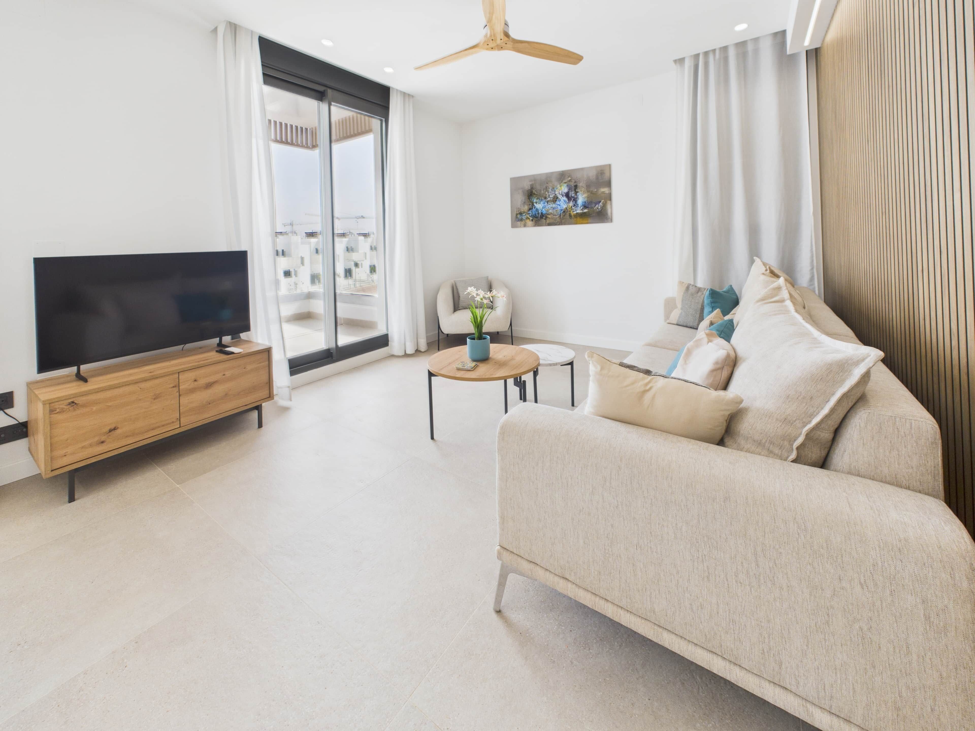 A modern living room with a flat-screen TV, wooden cabinet, sofa, coffee table, and a view of the outside through large windows.