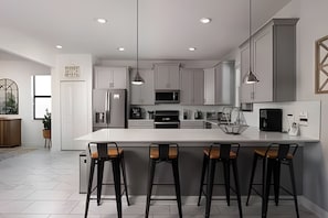 A modern kitchen with a large island, bar stools, stainless steel appliances, pendant lighting, and a view of the outdoors. - Luxury 6BR Villa w/ Pool, Spa & Near Disney (Kissimmee)