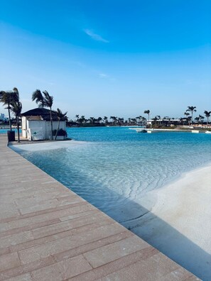Een helderblauw zwembad met een wit zandstrand, een klein gebouw met een bordje en palmbomen op de achtergrond.