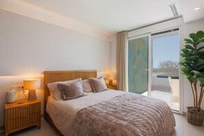 Une chambre à coucher avec un lit, des tables de chevet, une lampe, une plante en pot et une vue sur l’extérieur, visible à travers une porte vitrée.