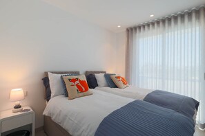 A double bed with a white and blue duvet, a nightstand with a lamp, and a window with sheer curtains. - Villa de los Suenos - Santa Rosalia Lake Resort (Torre-Pacheco)