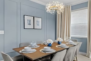 A modern dining room with a wooden table set for six, light blue walls, a chandelier, and a window with blinds. - 5BR Villa w/ Pool & Game Room Near Disney (Davenport)