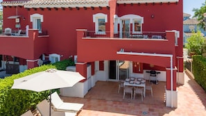 Edifici vermell de dues plantes amb un pati que inclou una taula, caires, un parasol blanc i una barbacoa.