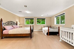 Een slaapkamer met een groot bed, een kleiner bed, een babybedje en een raam met uitzicht op groen.
