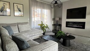 Un salon moderne avec un canapé gris, une table basse ronde, une plante en pot, une cheminée et deux œuvres d’art encadrées accrochées au mur.