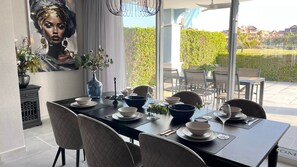 Un coin repas avec une table dressée pour six personnes, entourée de chaises, et offrant une vue sur un jardin et des bâtiments en arrière-plan.