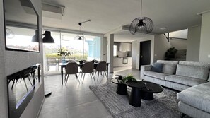 A modern living room with a grey sofa, a dining area with chairs and a table, and a kitchen in the background. - Villa Oliver - Mar Menor Golf Resort (Torre-Pacheco)