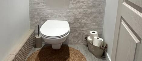 A bathroom with a toilet, a bidet, a toilet paper holder, a waste bin, and a woven rug on the floor.