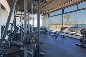Ett gym med utrustning för styrketräning, bänkar och en utsikt över palmer och en bil genom stora fönster.