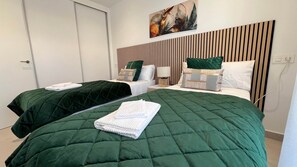 Une chambre d’hôtel avec deux lits, chacun recouvert d’une courtepointe matelassée verte et agrémenté de serviettes blanches. Un tableau est accroché au-dessus des lits, et les têtes de lit sont en lattes de bois.