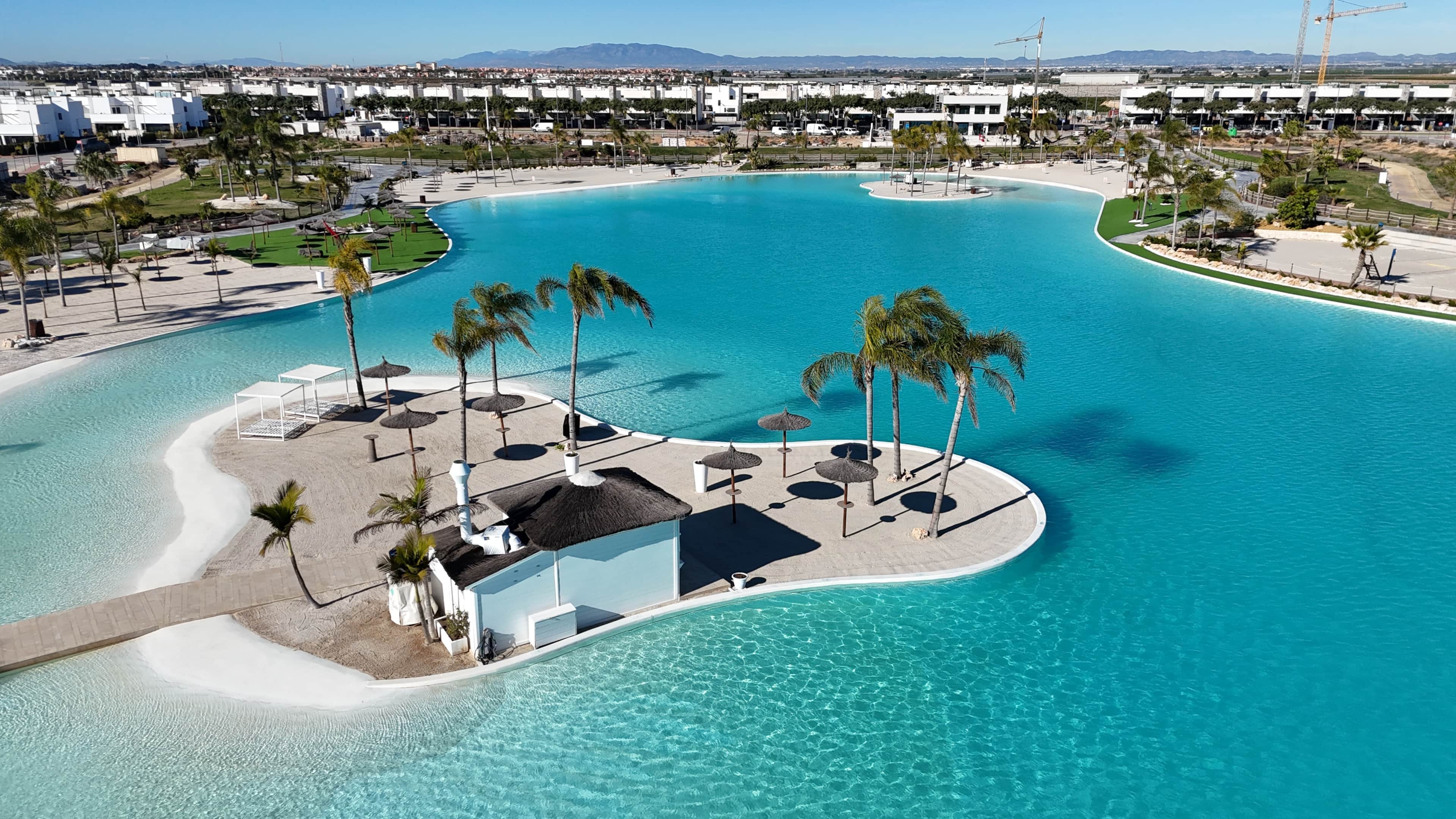 Vista aèria d'un complex turístic amb una gran piscina corbada envoltada de palmeres i gandules.