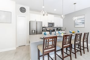 A modern kitchen with white cabinetry, a stainless steel refrigerator, a microwave, and a dining area with wooden chairs and a central island. - Champions Gate 4BR w/ Pool + Themed Rooms (Kissimmee)
