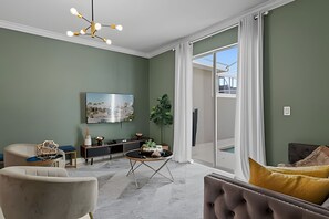 A living room with a modern chandelier, a flat-screen TV, a dining table with chairs, and a large window with curtains. - Disney-area 4BR Townhome w/ Heated Pool (Davenport)