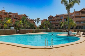 A swimming pool with a curved edge, surrounded by a low wall and palm trees. - La Casa Soleada - Mar Menor Golf Resort (Torre-Pacheco)