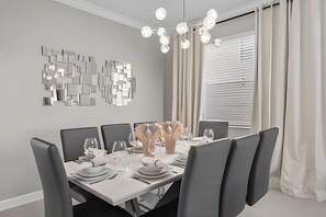 Un comedor moderno con una mesa rectangular dispuesta para seis personas, rodeada de sillas tapizadas en gris. Se observa una pieza decorativa en la pared y una araña con varias luces.