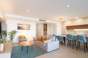 Ein modernes Wohnzimmer mit einer Couch, einem Couchtisch und Sesseln, ein Essbereich mit Tisch und Stühlen sowie eine Küche mit Arbeitsplatte und Barhocker.