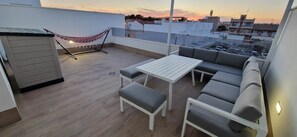Eine Dachterrasse mit einem weißen Tisch, einer grauen Couch, einer Hängematte und einem kleinen Schuppen.
