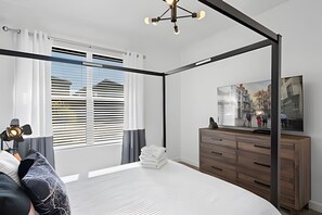 A modern bedroom with a four-poster bed, a large window with blinds, a wooden dresser, and a flat-screen TV on the wall. - 5BR Home w/ Pool, Game Room + Near Disney (Davenport)