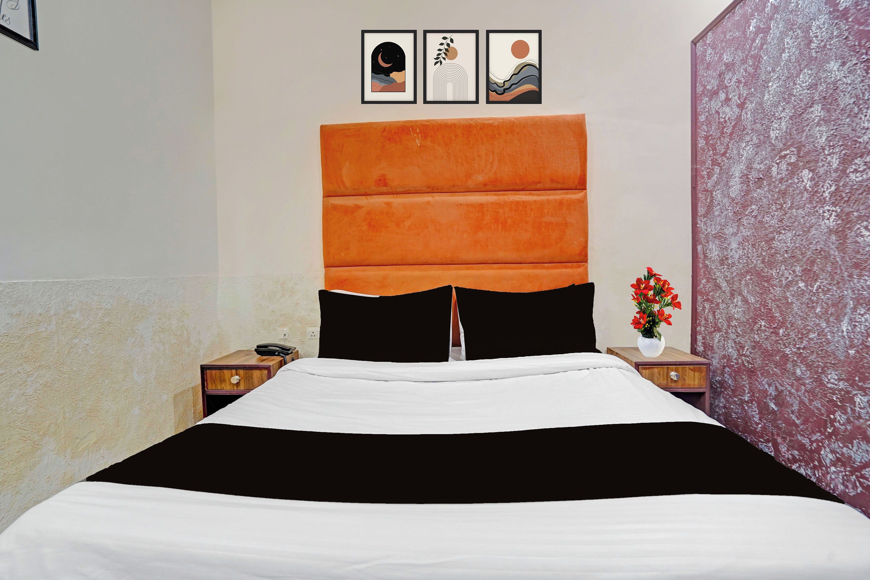 A bed with a black and white bedspread, an orange headboard, and a nightstand with a phone and a vase of flowers.