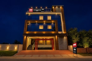 Um hotel moderno com estrutura em dois andares, entrada destacada com moldura vermelha e sinalização indicando disponibilidade de quartos no aplicativo.