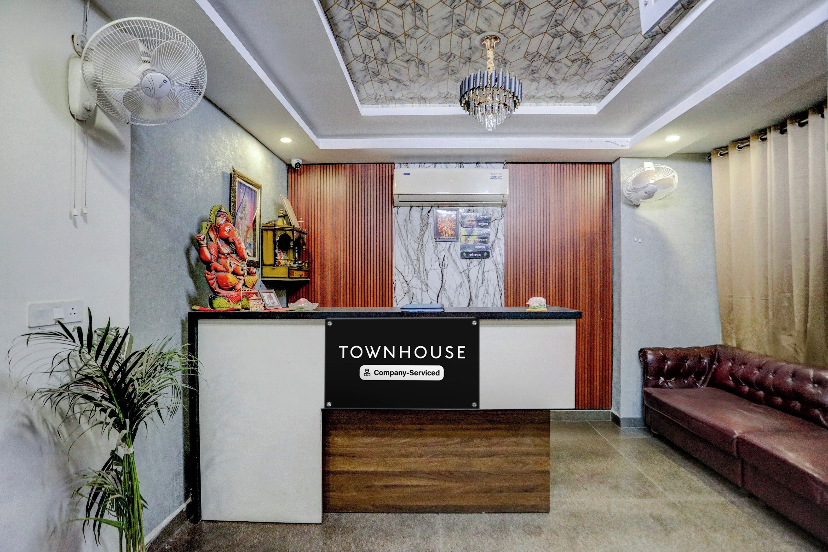Ein Empfangstresen mit dem Schild 'TOWNHOUSE Company-Serviced', ein Deckenventilator, eine Kristallleuchter, ein braunes Ledersofa und ein Topfpflanze.