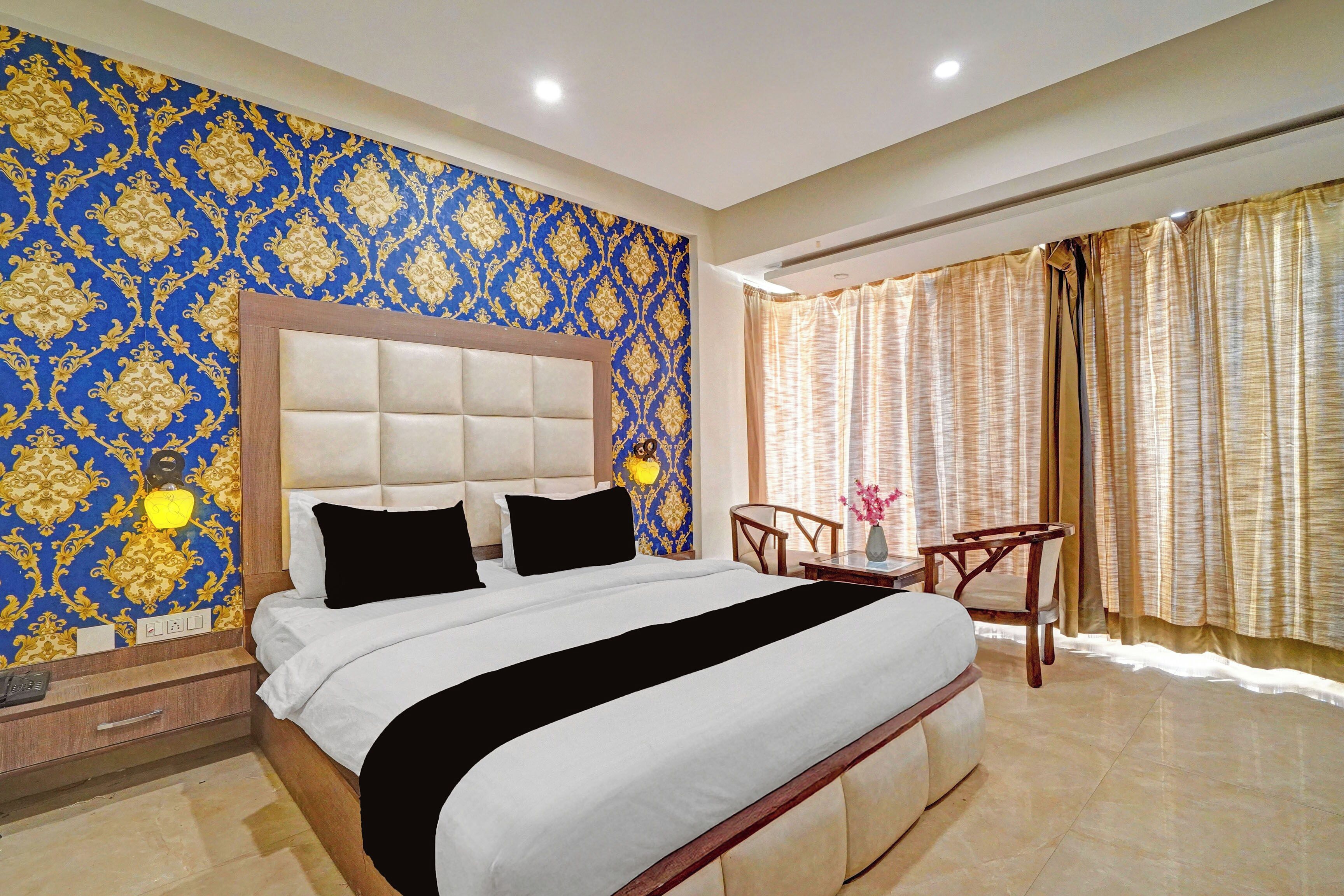 Habitación de hotel con una cama grande, cabecero de madera, pared con estampado, ventana con cortinas y una pequeña mesa con sillas.