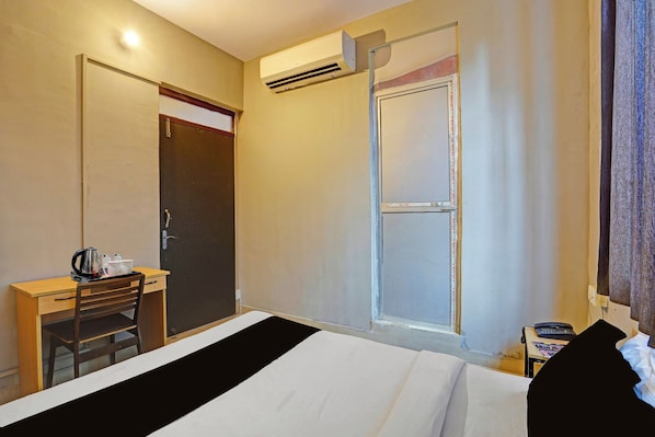 Quarto de hotel com uma cama, uma escrivaninha com chaleira, uma cadeira, uma porta e um aparelho de ar condicionado.