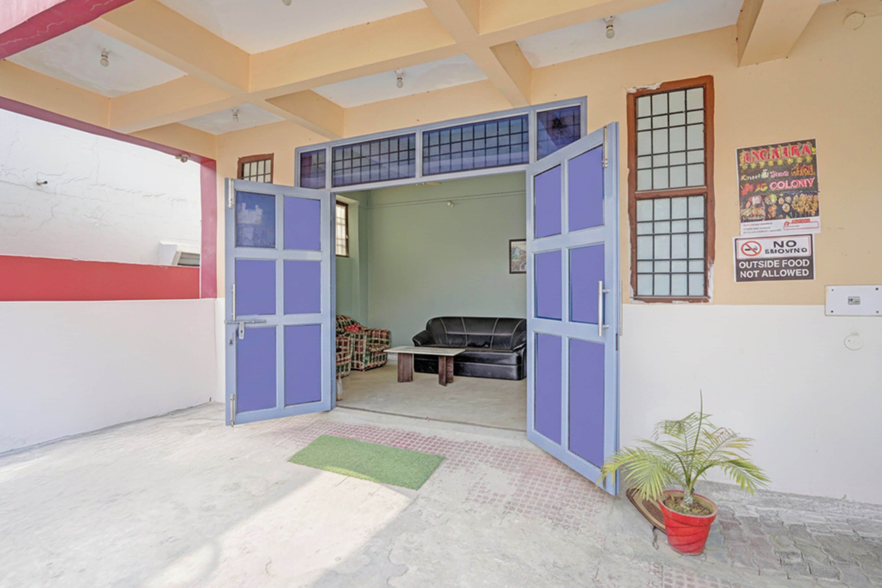 Un umbral abierto que da a una habitación con un sofá, una mesita pequeña, una planta en maceta y una pared con un letrero que dice 'No se permite comida de fuera'.