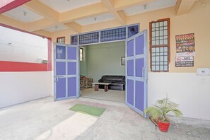 Een open deur die leidt naar een kamer met een bank, een kleine tafel, een potplant en een muur met een bord waarop staat 'Geen eten van buiten'.