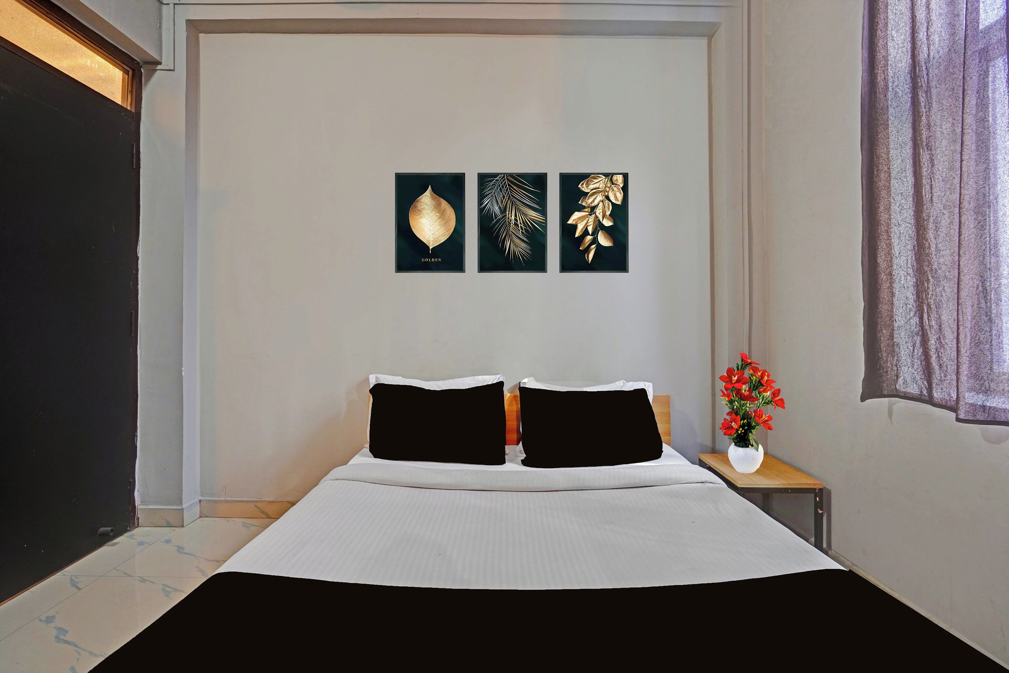 Un dormitorio con una cama, dos almohadas negras, una mesita de noche blanca con un florero de flores rojas y tres cuadros decorativos en la pared.