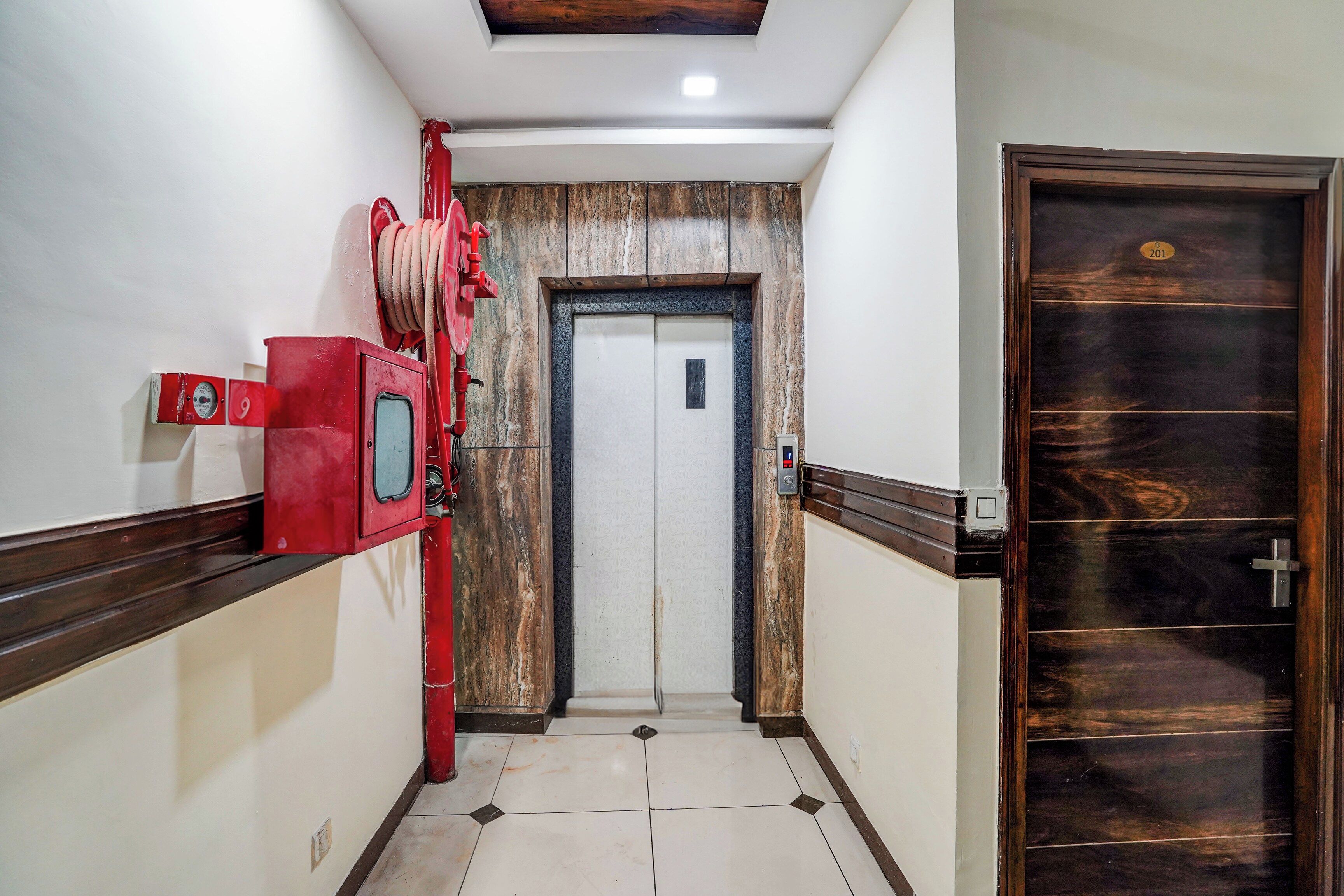 Un pasillo con un detector de incendios rojo, una puerta de madera con placa de número y suelo de baldosas.