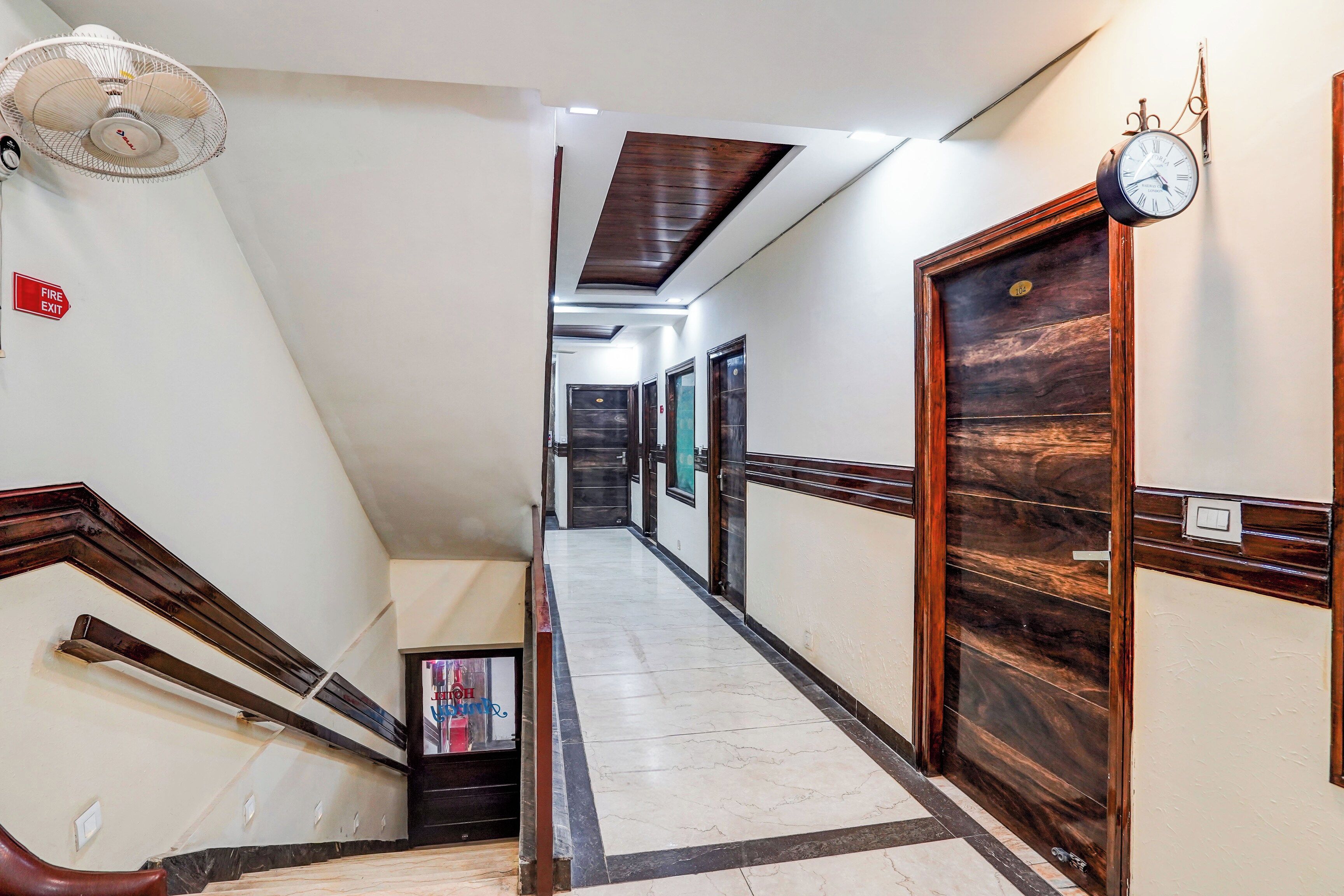 Pasillo de hotel con una escalera, señal de salida de incendios, un reloj y una puerta con placa de número.