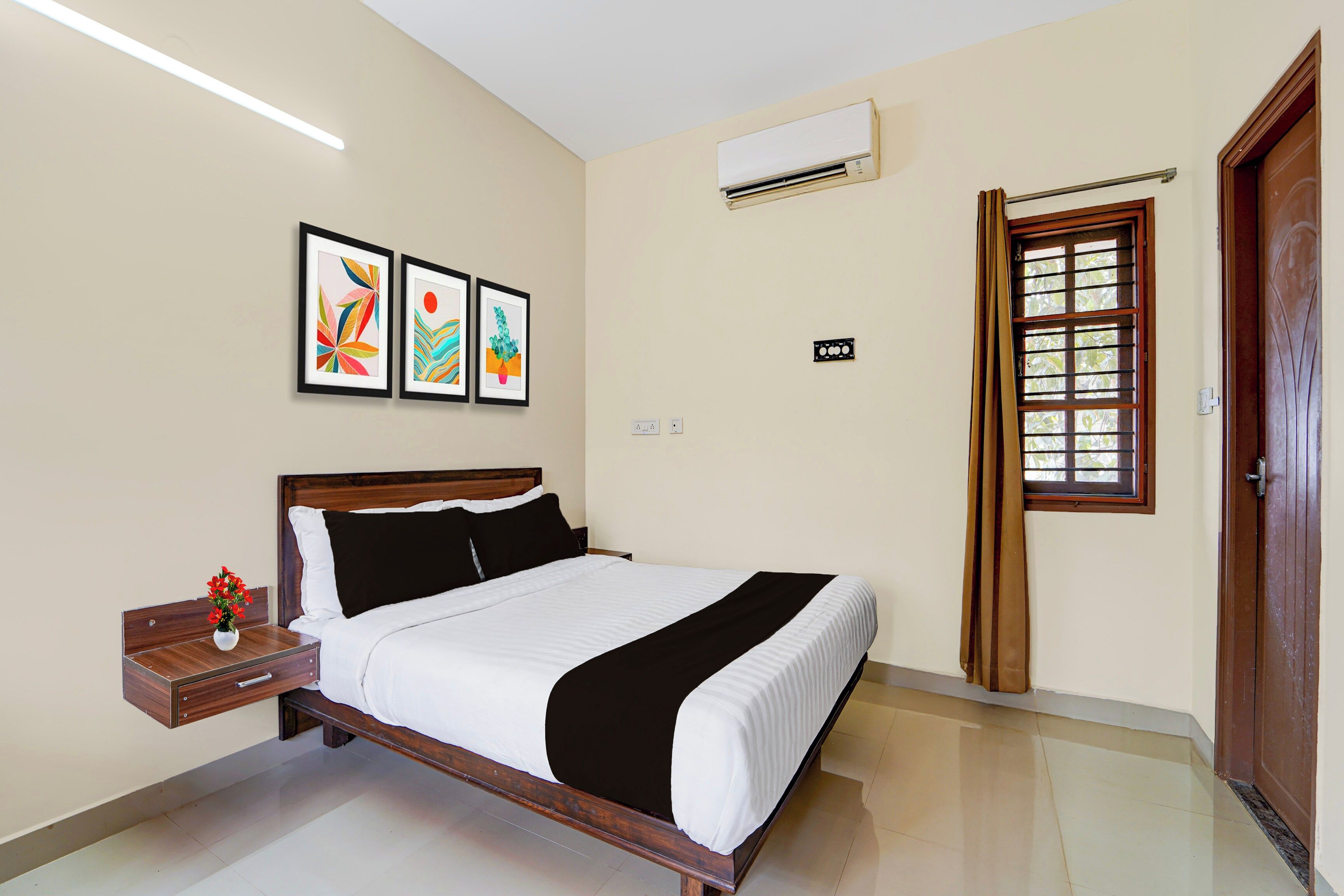 A hotel room with a bed, a nightstand with a vase of flowers, a wall-mounted air conditioner, a window with a curtain, and three framed artworks on the wall.
