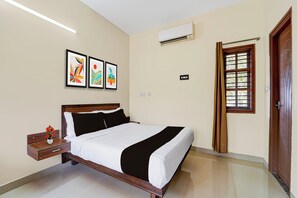 A hotel room with a bed, a nightstand with a vase of flowers, a wall-mounted air conditioner, a window with a curtain, and three framed artworks on the wall.