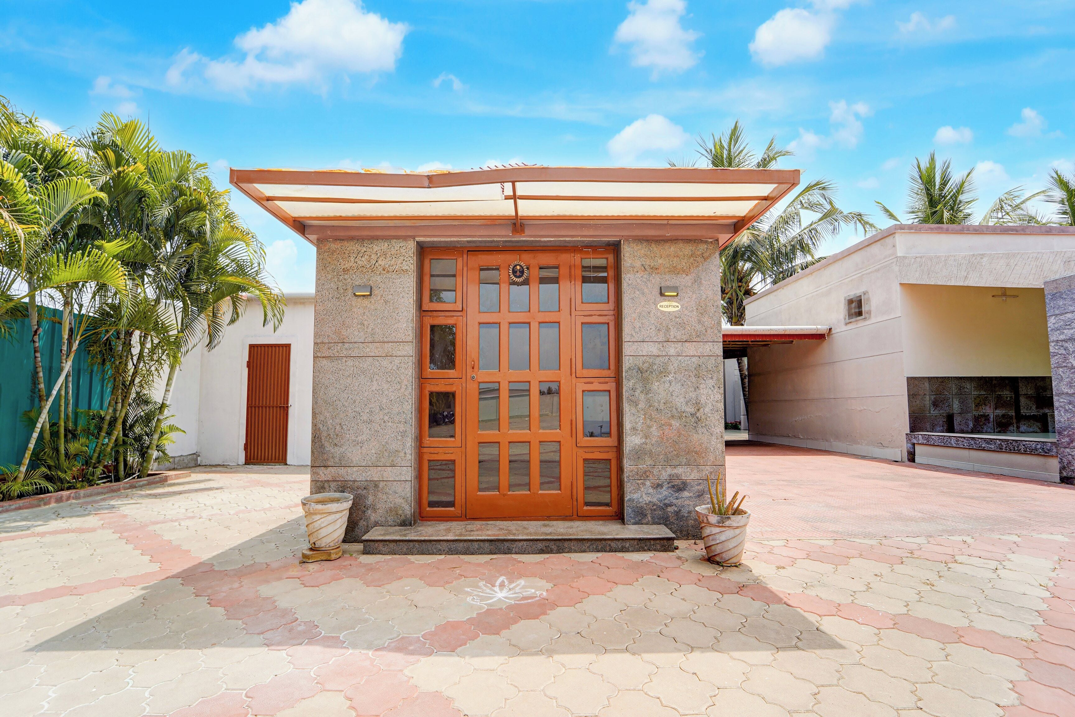 A building with a central entrance featuring a wooden door with glass panels, a reception area, and a paved courtyard with palm trees.
