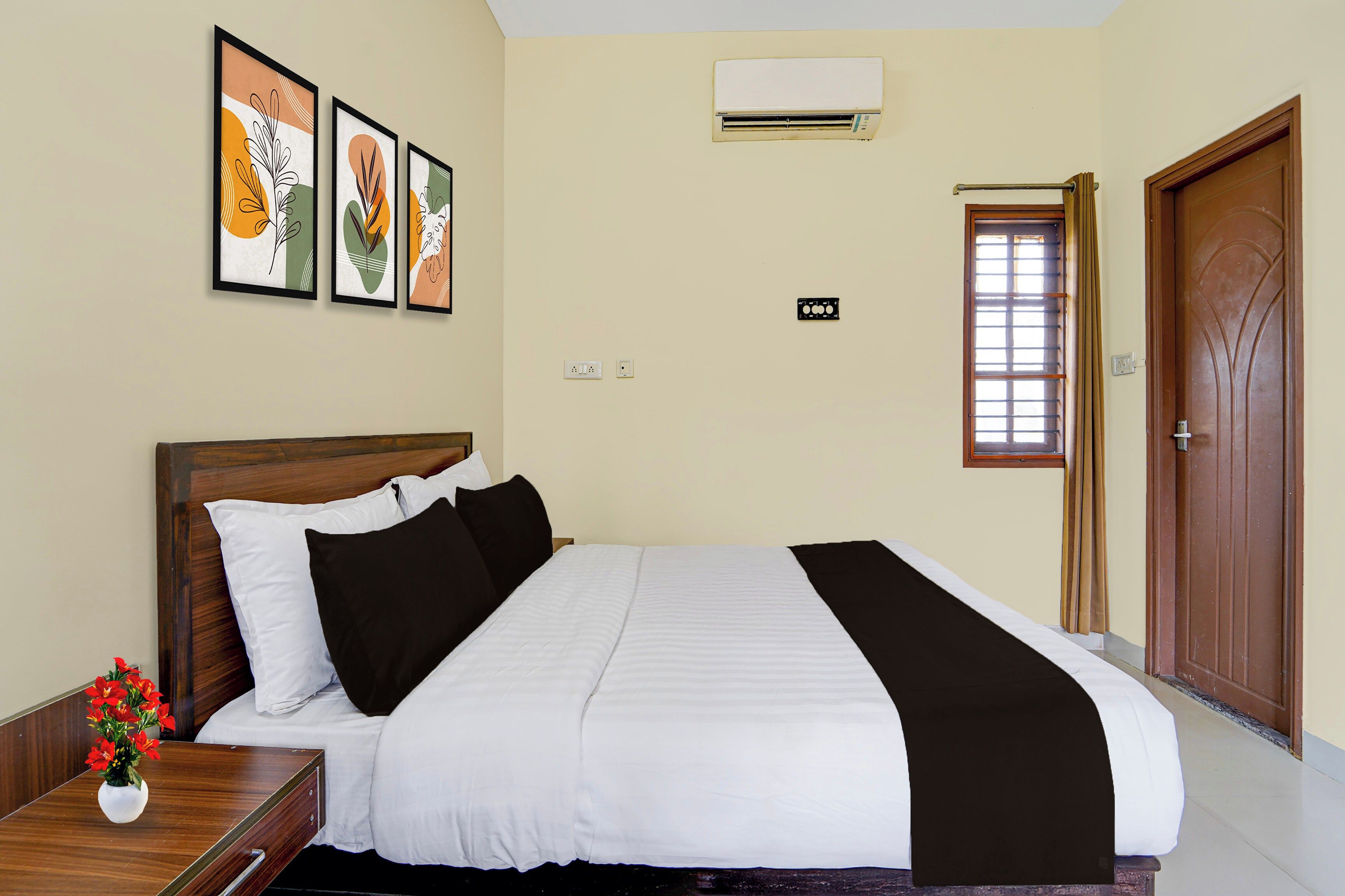 Habitación de hotel con cama, mesita de noche, un jarrón de flores, ventana con persianas y puerta.