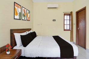 Habitación de hotel con cama, mesita de noche, un jarrón de flores, ventana con persianas y puerta.