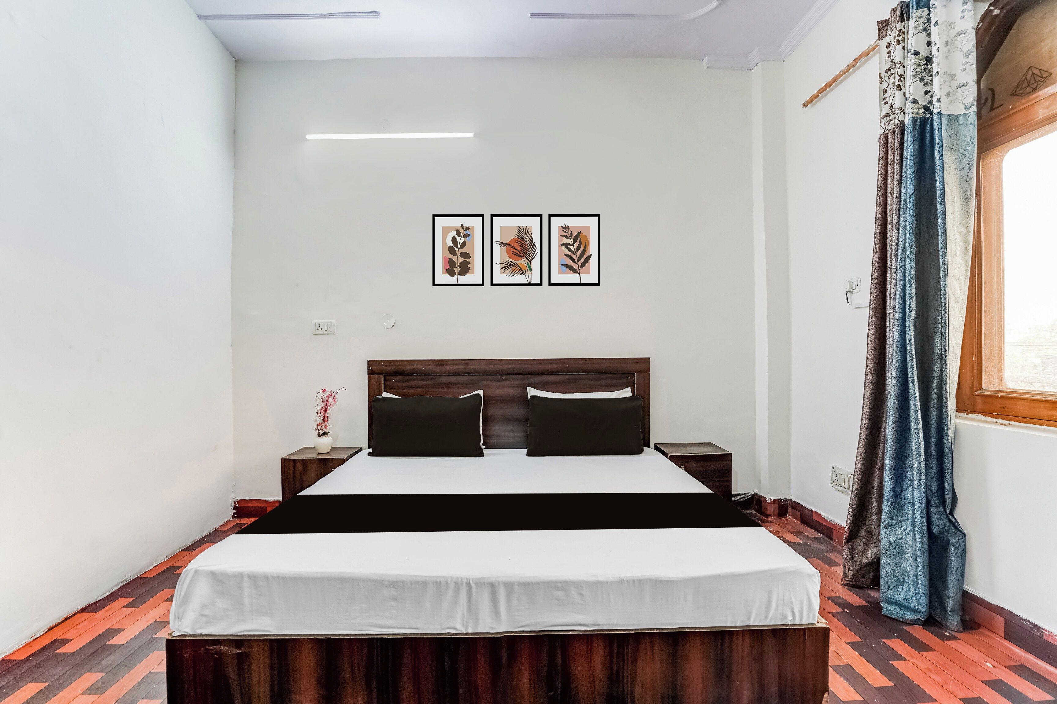 A bedroom with a wooden bed, white bedding, a nightstand, a vase with flowers, and a window with curtains.