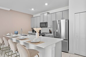A modern kitchen with white cabinetry, a central island with seating, a stainless steel refrigerator, and a microwave. - Disney Area Championsgate 4BR Townhome w/ Pool (Davenport)