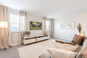 A modern living room with a white sofa, a flat-screen TV mounted on the wall, a white console table, and a large window with curtains. - 6BR Villa w/ Pool & Game Room Near Disney (Davenport)
