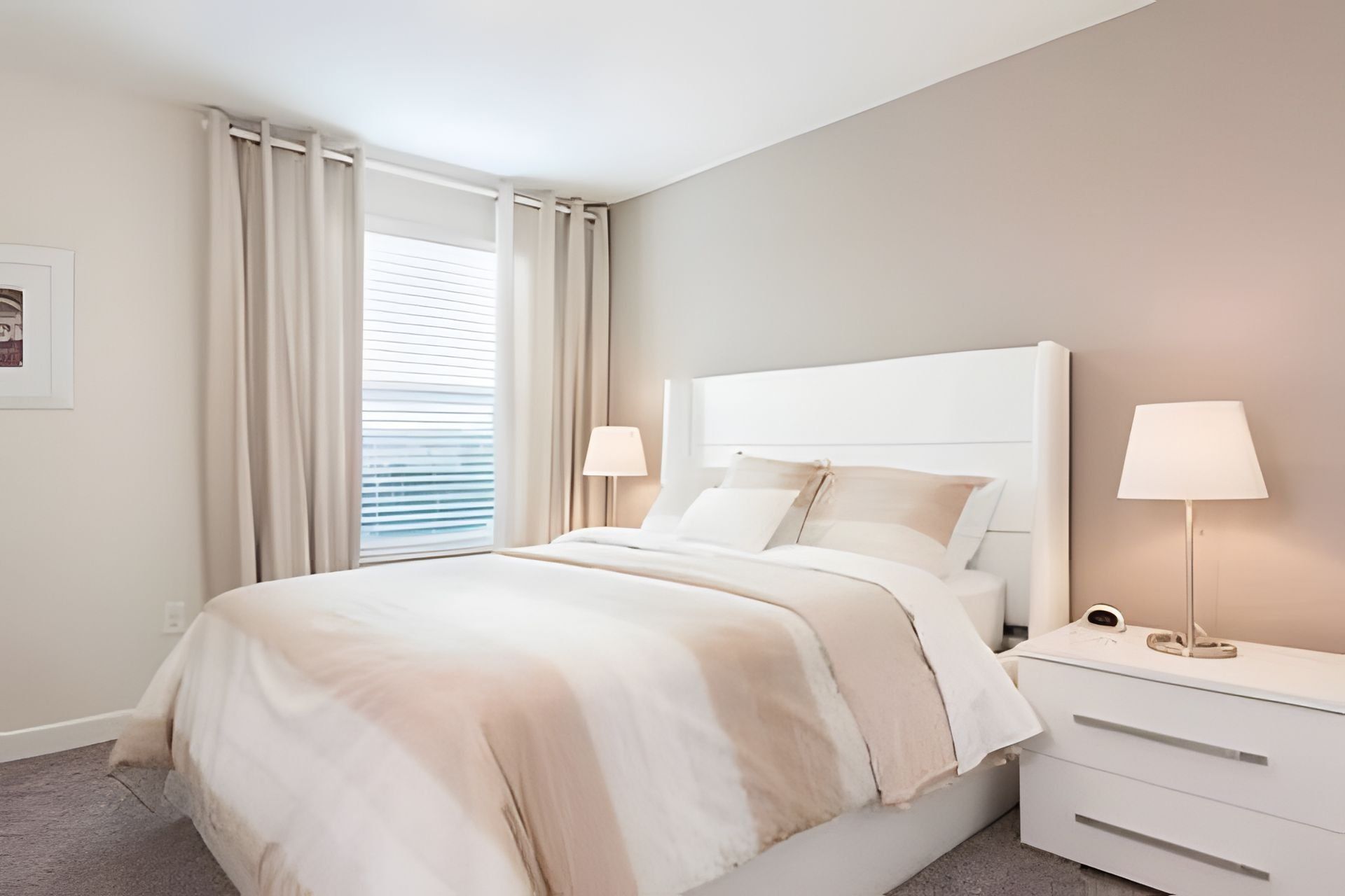 A modern bedroom with a large bed, two bedside tables with lamps, a window with curtains, and a framed picture on the wall.
