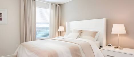 A modern bedroom with a large bed, two bedside tables with lamps, a window with curtains, and a framed picture on the wall.
