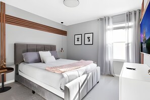 A modern bedroom with a large bed, a bedside table, a desk, and a window with curtains. - Luxury 7BR Villa w/ Pool & Game Room (Kissimmee)