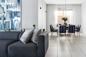 A modern living room with a dark grey sofa, a glass coffee table, and a dining area with a white table, dark chairs, and a potted plant. - Luxury 7BR Villa w/ Pool & Game Room (Kissimmee)