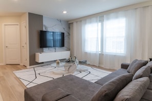 A modern living room with a grey sofa, a glass coffee table, a flat-screen TV mounted on a marble wall, and a large window with sheer curtains. - Windsor Cay 5BR Villa w/ Pool & Game Room (Clermont)