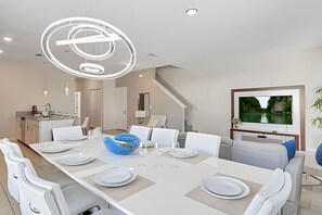 Un comedor moderno con una mesa larga, sillas blancas, un cuenco decorativo azul y un televisor de pantalla plana en la pared.