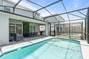 A covered outdoor swimming pool with a glass enclosure, patio seating, and a view of a house in the background. - Luxury 5BR Villa With Pool and Themed Bedrooms (Clermont)
